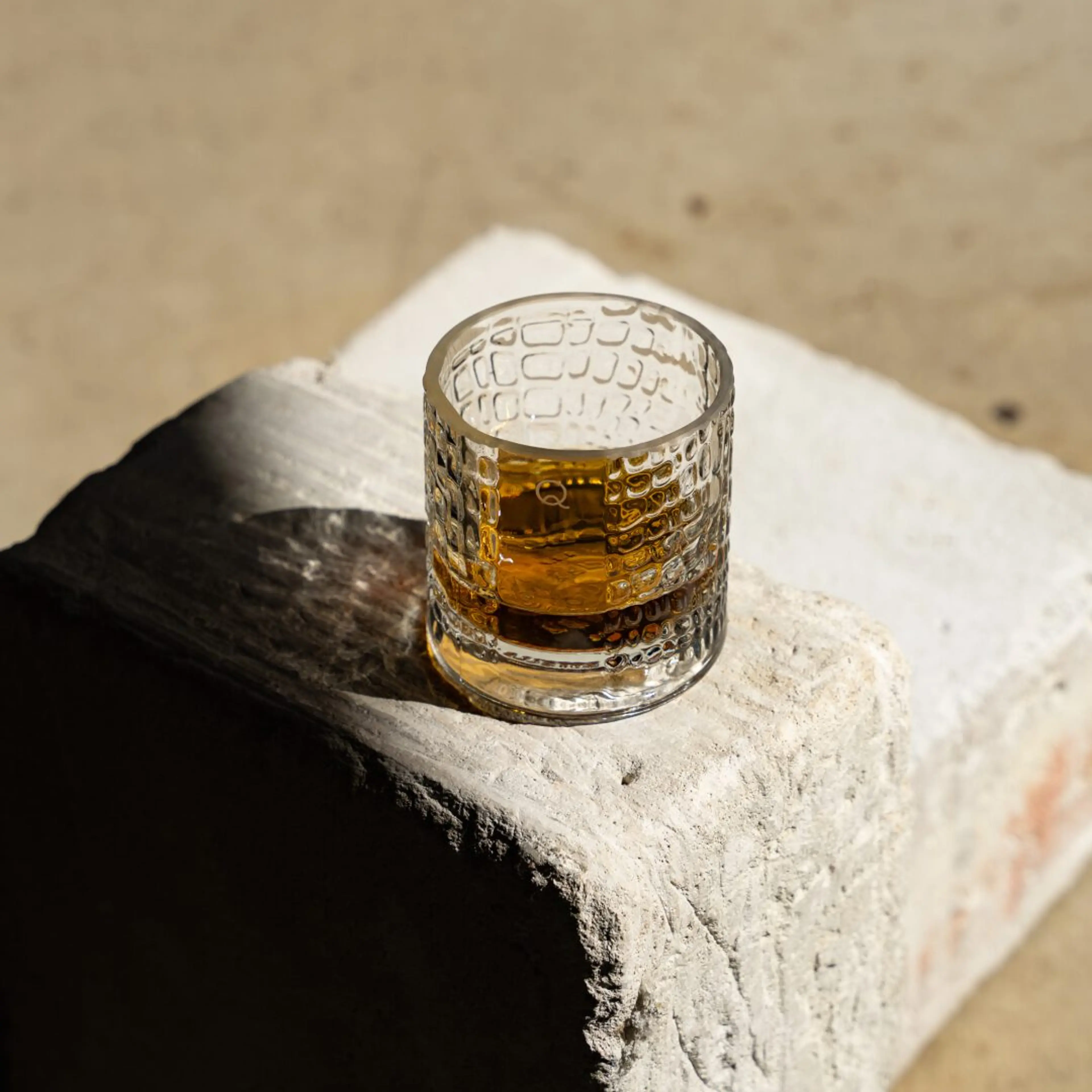 Textured glass tumbler with amber liquid on a rough white stone block in sunlight