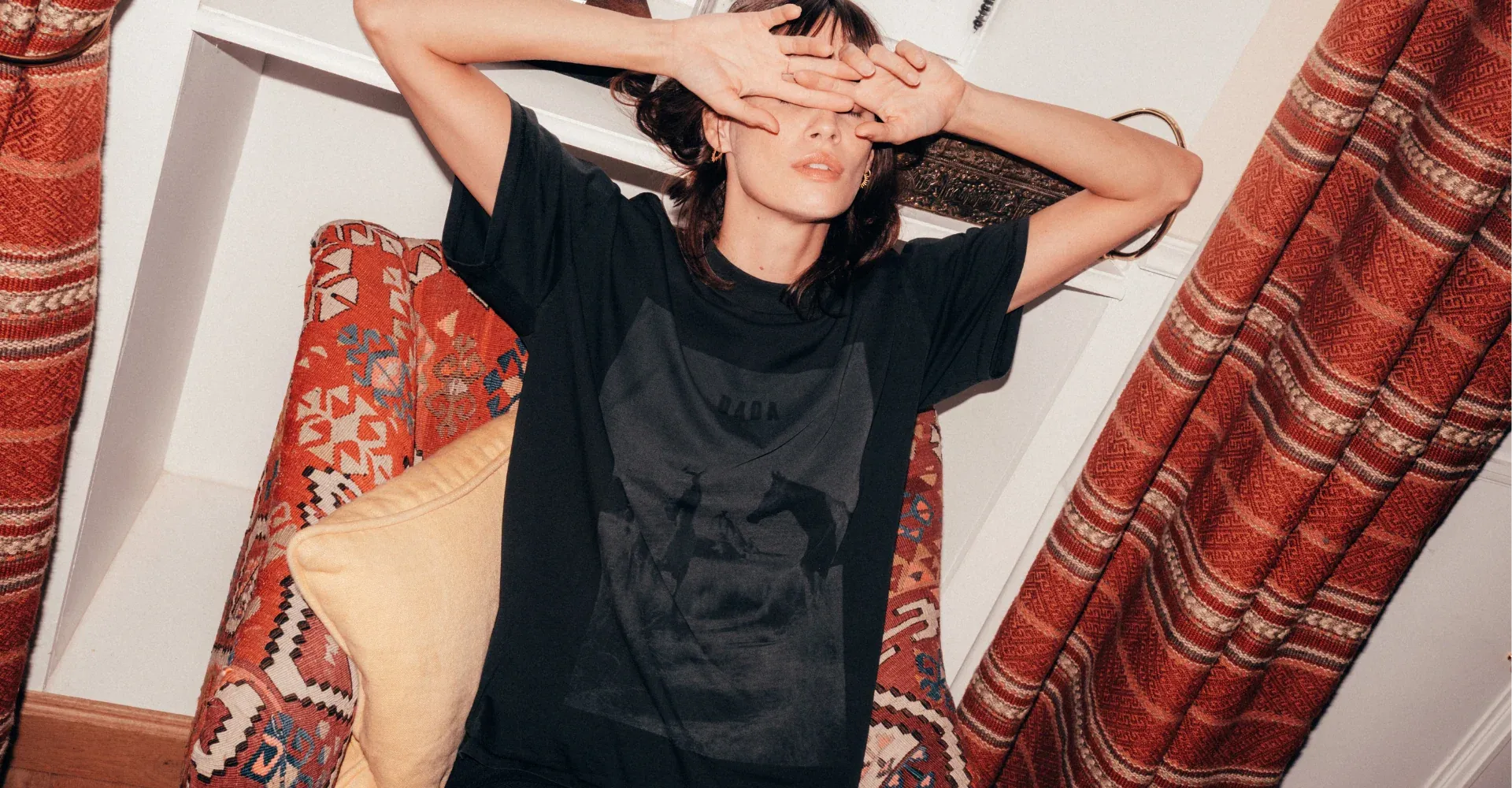 Woman in a black horse-print T-shirt covers her eyes with her hands on a kilim chair by red curtains