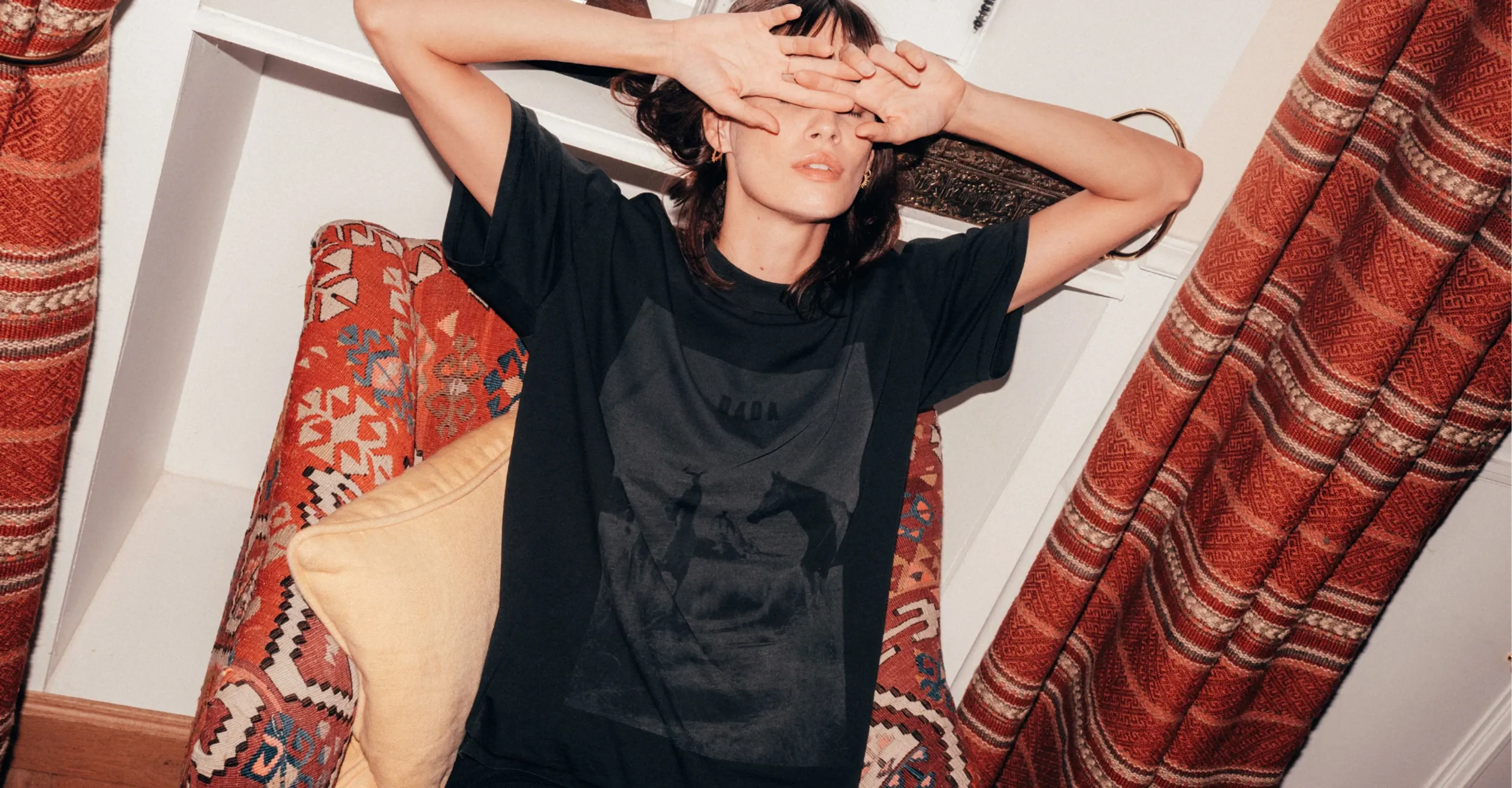 Woman in a black horse-print T-shirt covers her eyes with her hands on a kilim chair by red curtains