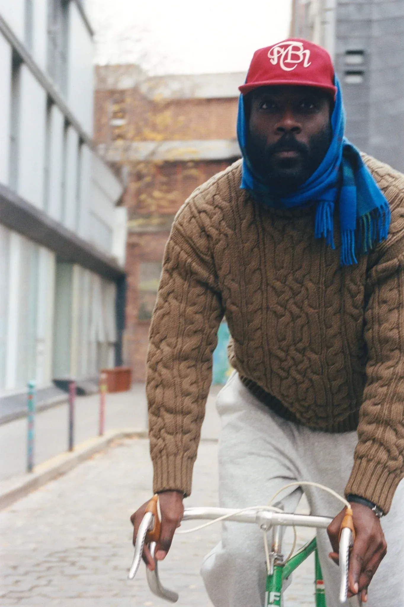 Man riding a bike on a cobbled street in a brown cable-knit sweater, blue scarf over his head and red cap