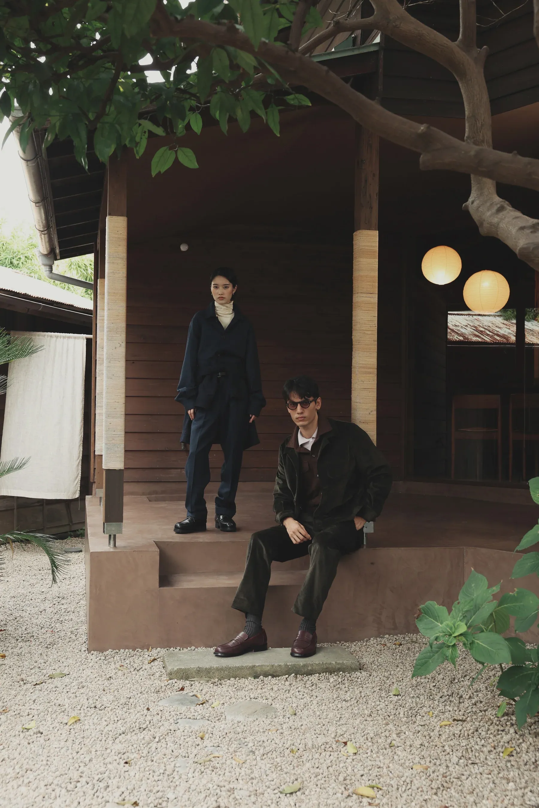 Woman in navy standing and man in dark corduroy seated on the porch of a wooden house
